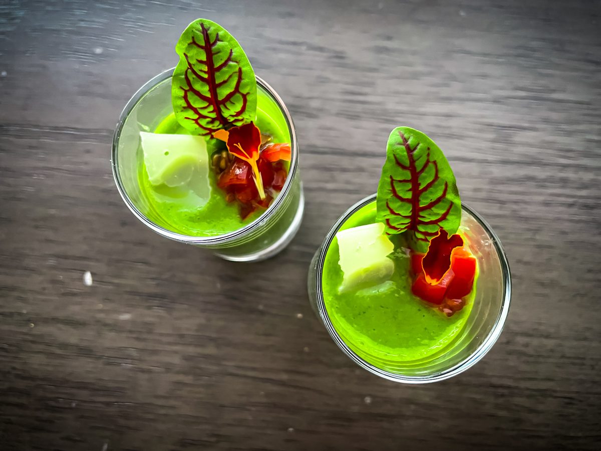 Two small glasses of bright green gazpacho topped with cucumber cubes, red pepper and a carved red leaf garnish, on a dark wood ta