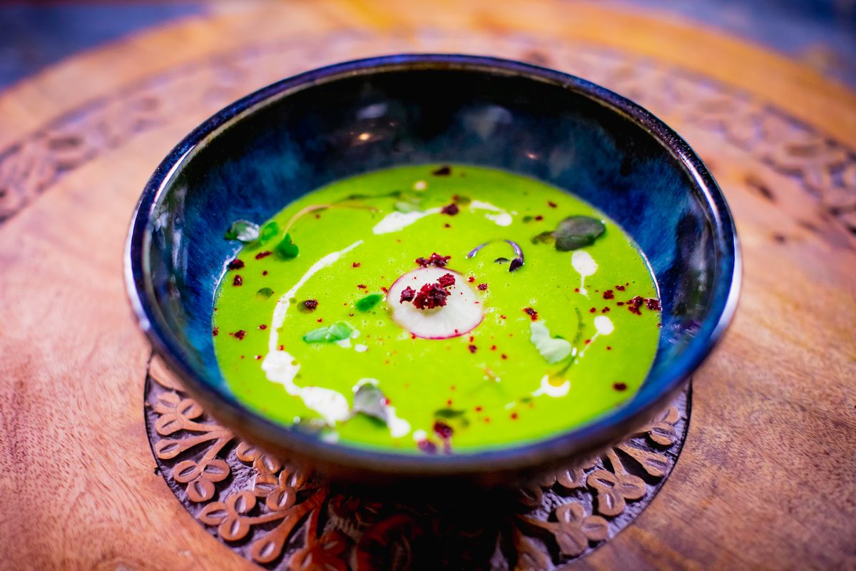 A bowl of smooth bright green pea soup swirled with cream and oil and dotted with herbs, in a blue glazed bowl on a carved dark wo