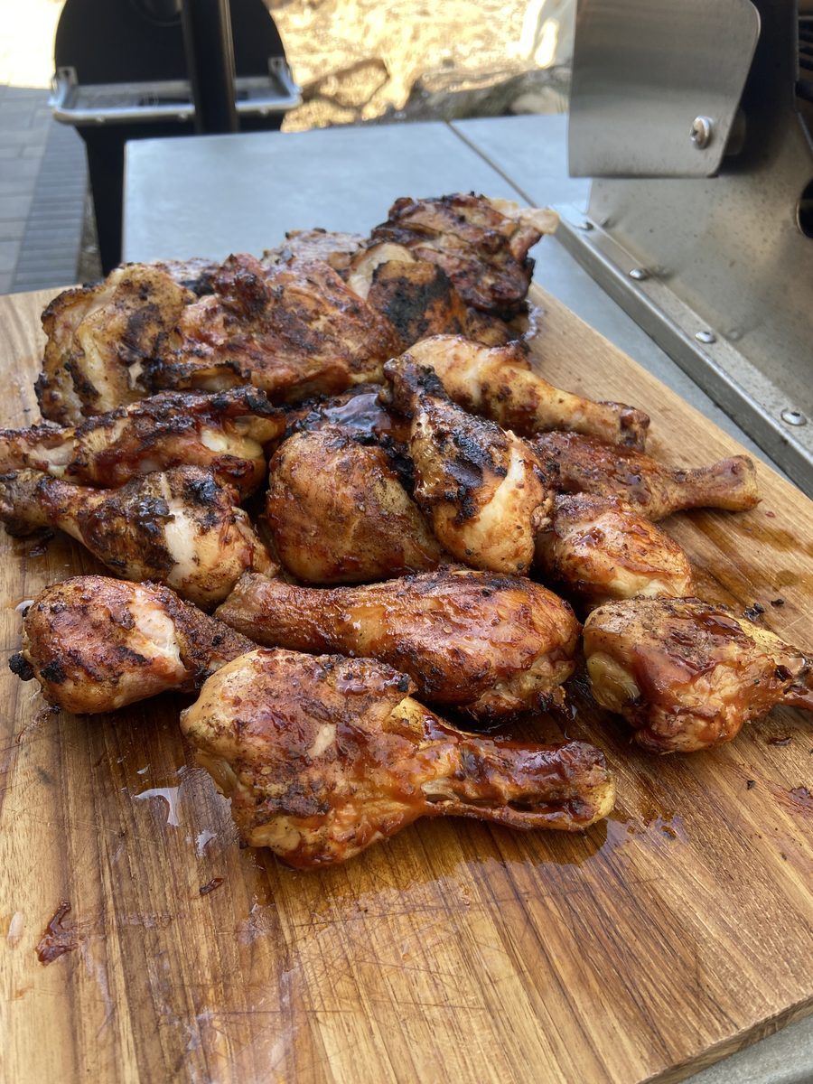 A heap of charred grilled chicken drumsticks and thighs piled on a wooden cutting board next to an outdoor grill, background clutt
