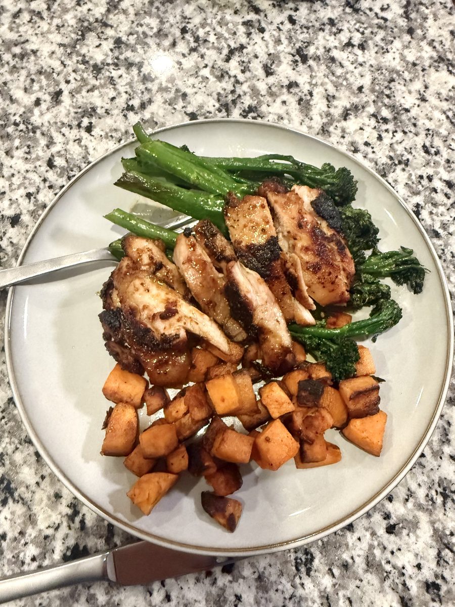 Sliced grilled chicken with charred broccolini and cubed roasted sweet potato arranged on a white plate on a speckled granite coun