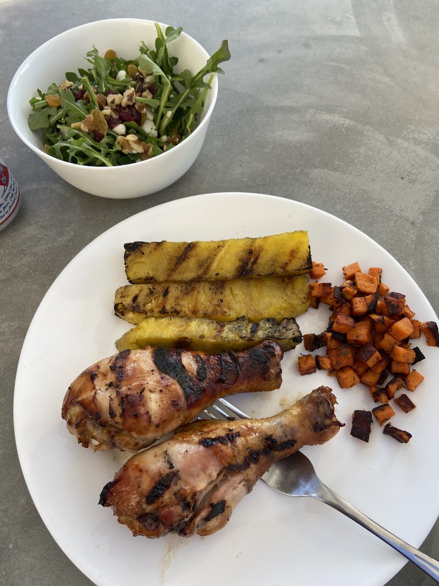 Overhead white plate of grilled chicken, grilled pineapple spears and diced roasted sweet potato with a side bowl of arugula salad