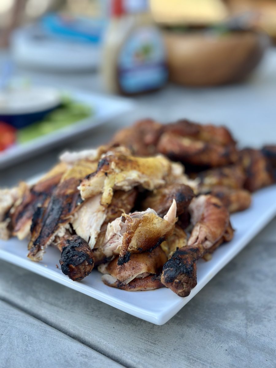 A white platter piled with charred grilled bone-in chicken pieces on an outdoor table with condiments in the background.