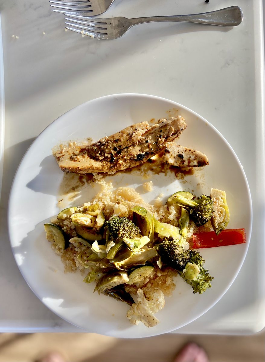 Sliced seasoned chicken breast with roasted broccoli, zucchini and cauliflower over quinoa on a white plate in harsh direct sunlig