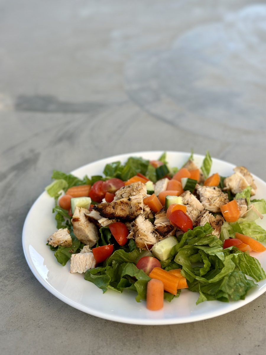 Chopped grilled chicken over romaine with cherry tomatoes, carrots and cucumber on a white round plate set on a grey stone counter
