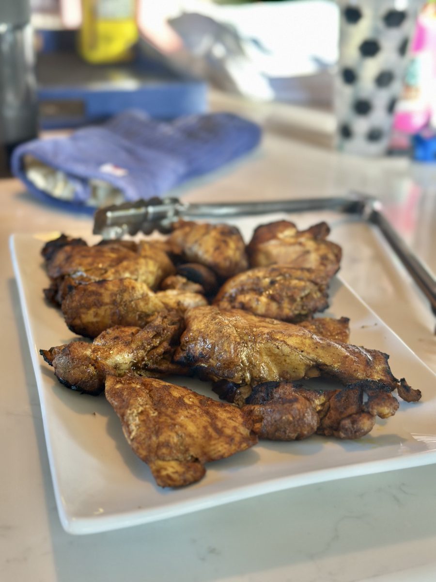 Grilled seasoned chicken thighs and wings piled on a white rectangular platter with metal tongs, on a kitchen counter with clutter