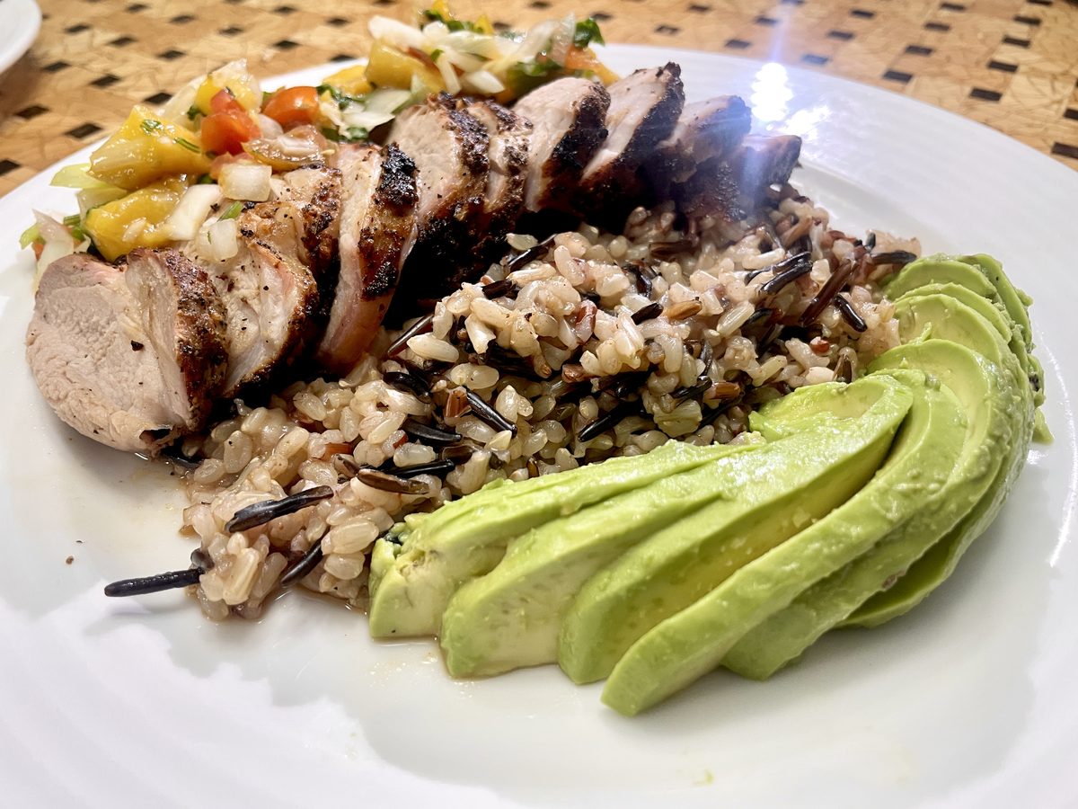 Sliced grilled chicken breast with wild rice, fanned avocado slices and a spoonful of mango salsa on a white plate on a woven plac