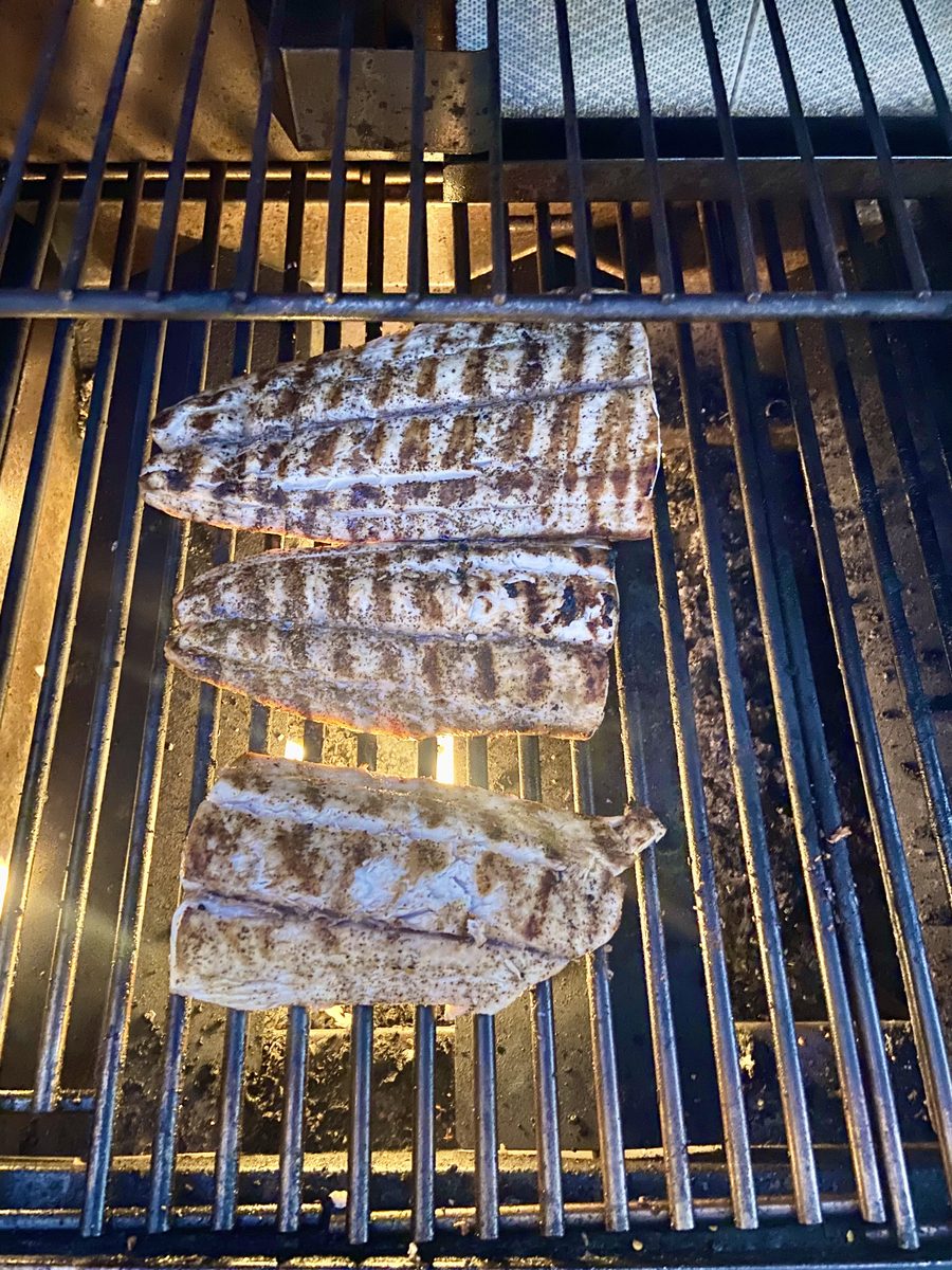 Several pale fish fillets laid across the blue-tinted grates of a hot grill, cooking with no people in frame