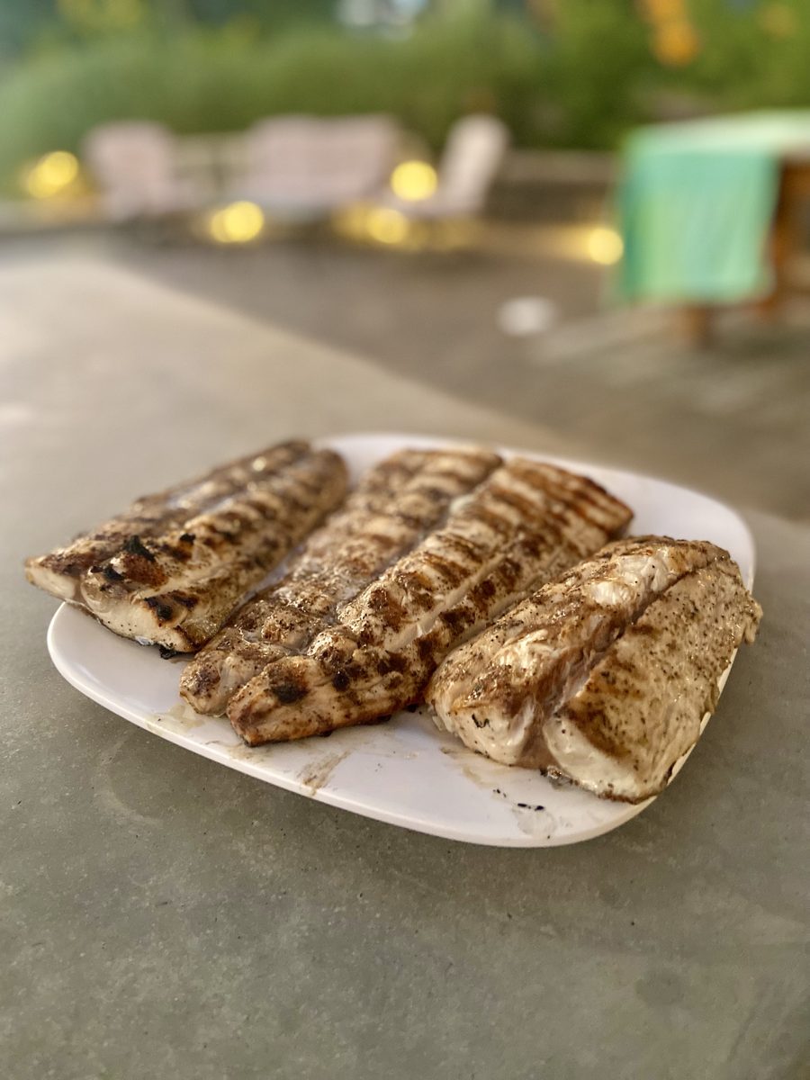 A white platter on an outdoor counter holding several grill-marked pale fillets; the flaky white flesh reads as fish, though grill
