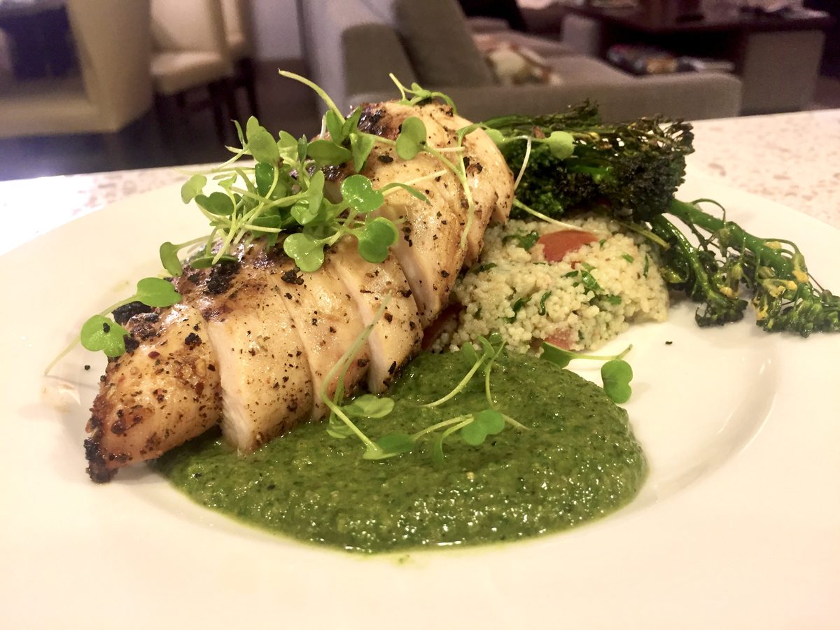 A grilled white fish fillet over a pool of green pesto sauce with couscous, broccolini and microgreens on a white plate in a brigh