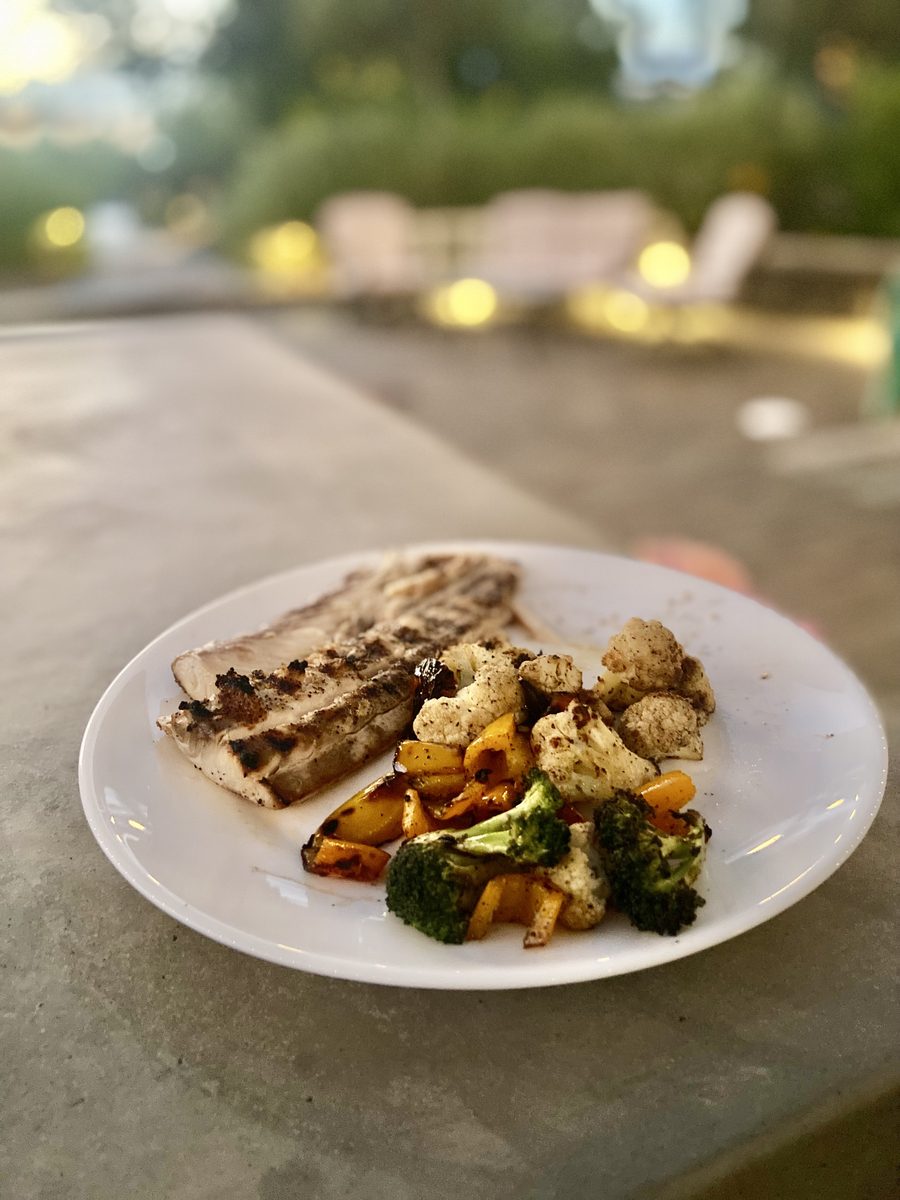 Grilled fish fillet with roasted cauliflower, broccoli and sweet potato on a white plate on an outdoor counter with warm bokeh lig