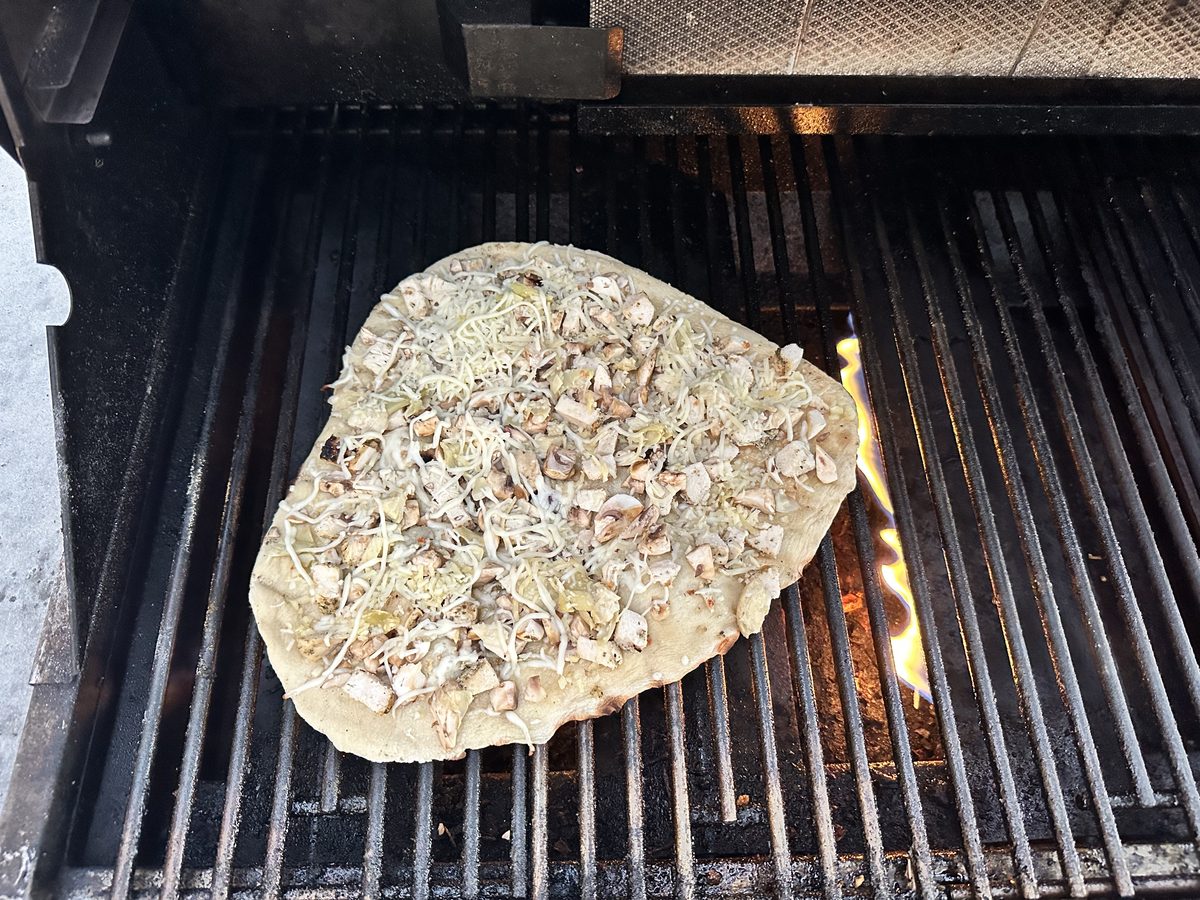 A rustic flatbread topped with shredded cheese, mushrooms and herbs cooking directly on gas grill grates with a flame visible besi
