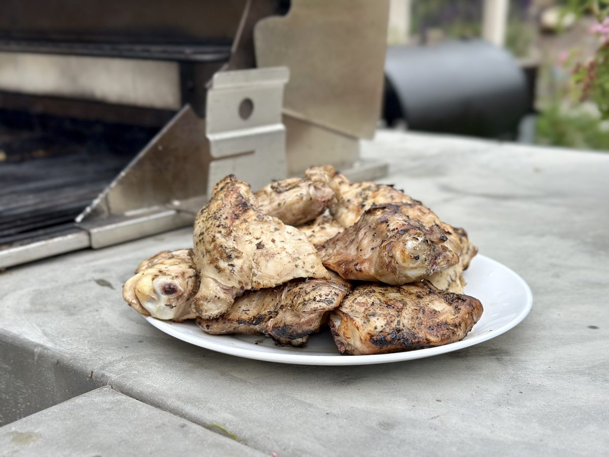 A pile of herb-seasoned grilled chicken leg quarters stacked on a white oval platter set on an outdoor concrete counter beside a g