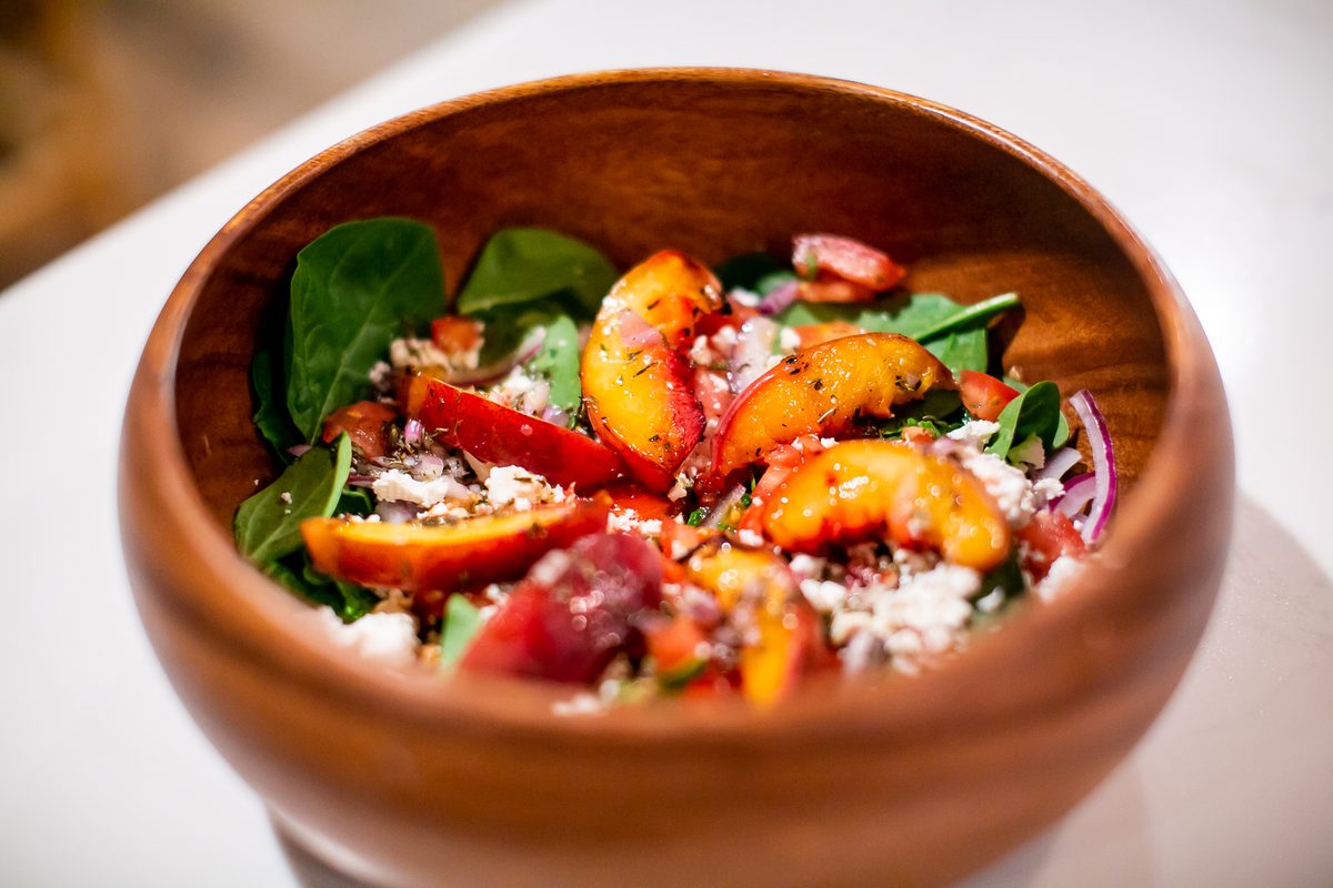 A wooden bowl holding a spinach salad with peach slices, red onion, tomato and crumbled white cheese.