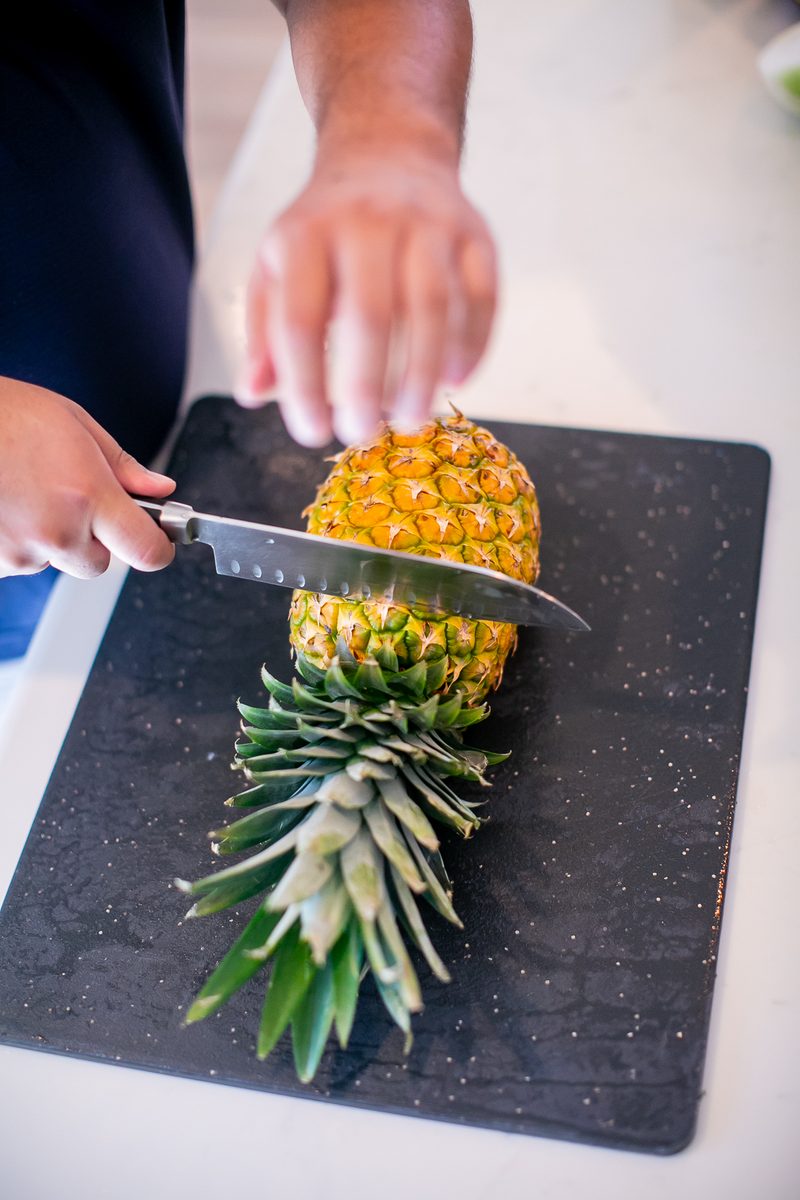A person in a navy shirt (torso only, no face) slicing the top from a whole pineapple with a chef's knife on a black cutting board