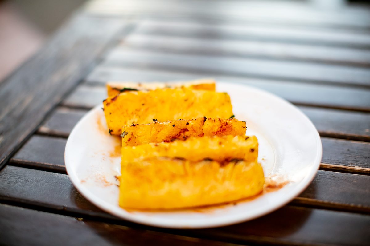A small white plate set on a grill grate holding two thick planks of grilled pineapple with dark char marks and a red sauce drizzl