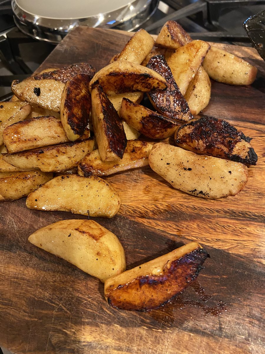 A pile of charred grilled potato wedges on a dark wooden cutting board set on a stovetop with a pan behind
