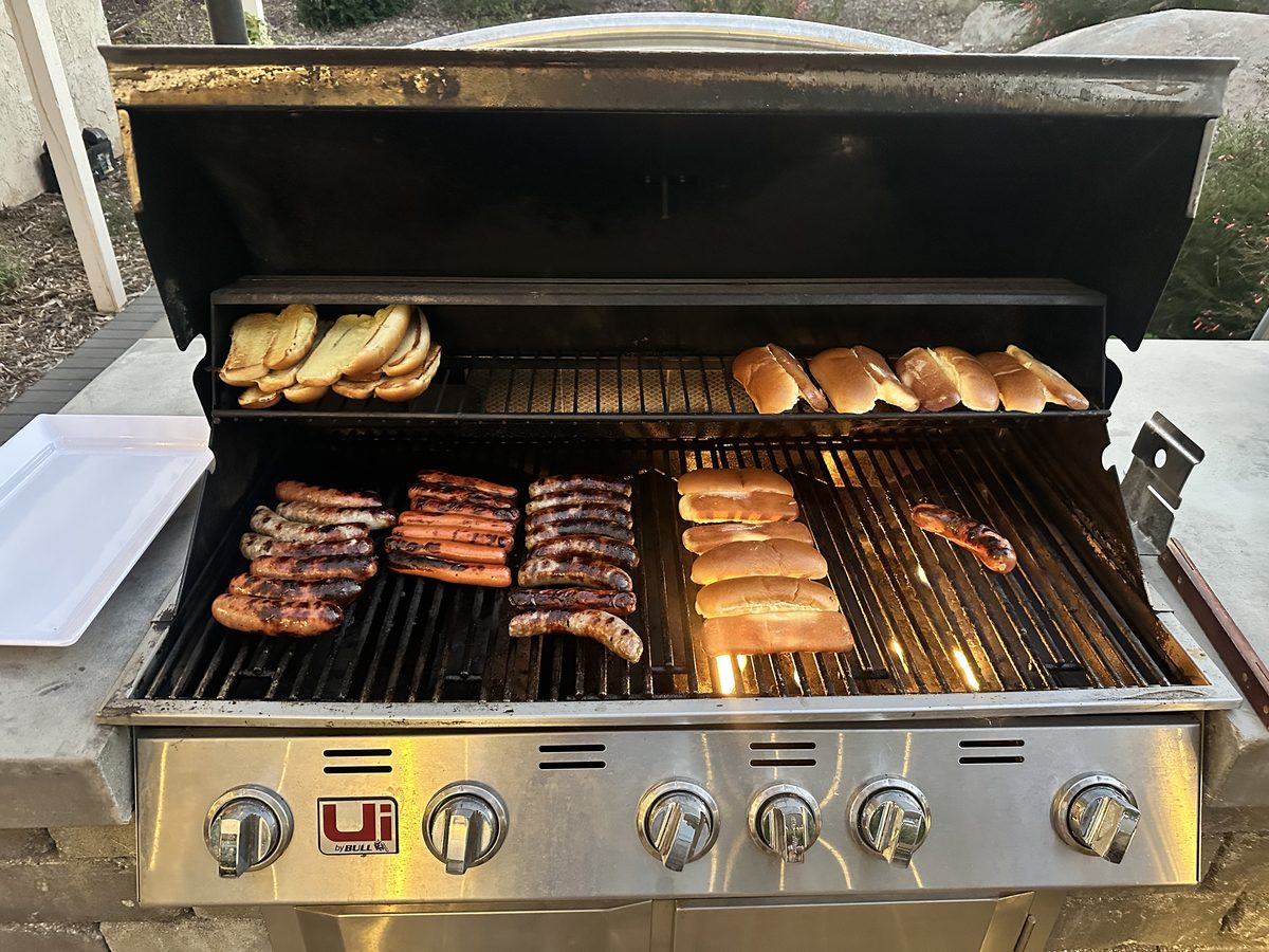 Sausages, ribs and hot dog buns toasting on the grates of an open stainless gas grill outdoors, no people in frame