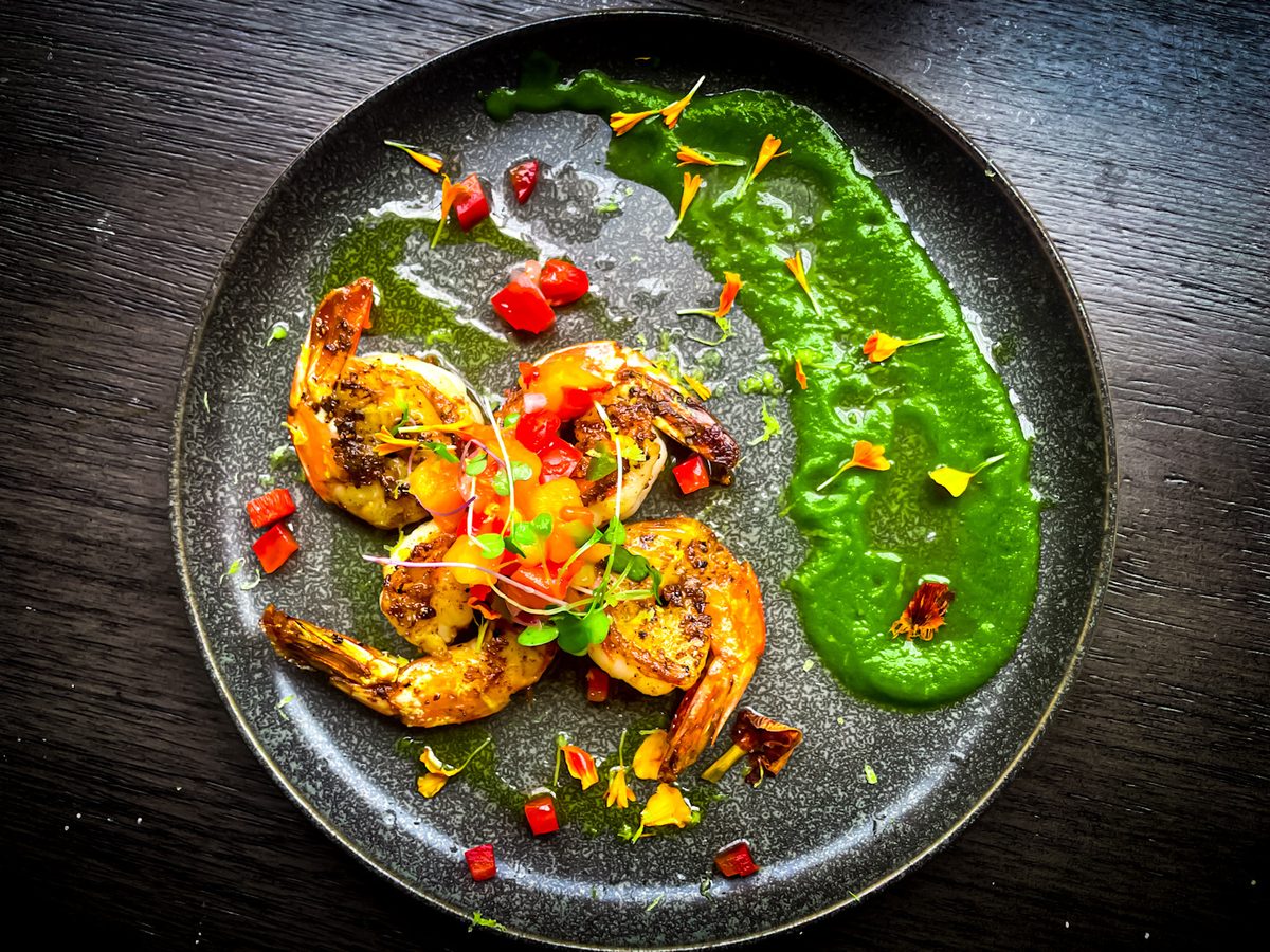 Grilled shrimp arranged over a smear of bright green chimichurri with cherry tomatoes and edible flower petals on a dark speckled
