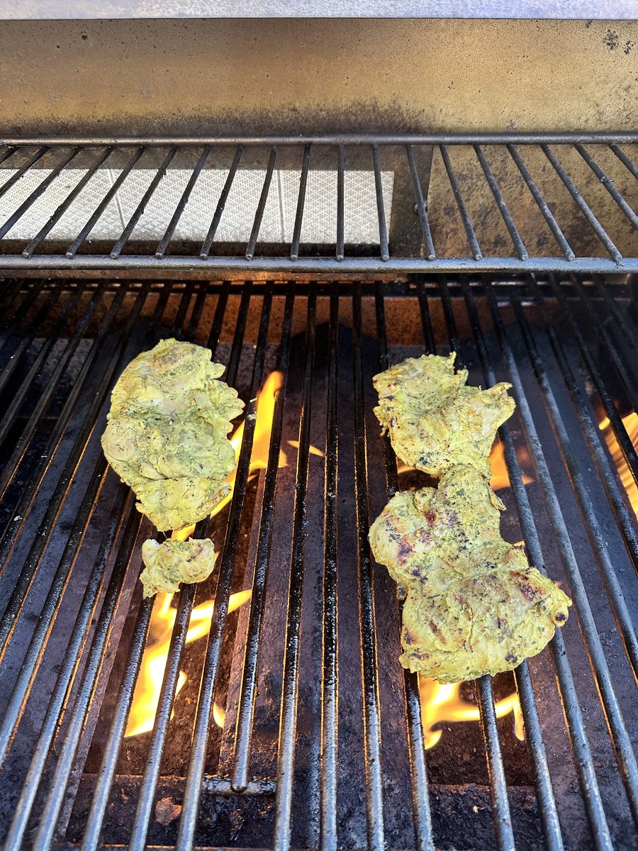 Two herb-marinated chicken thighs cooking on the grates of an outdoor grill, no people in frame