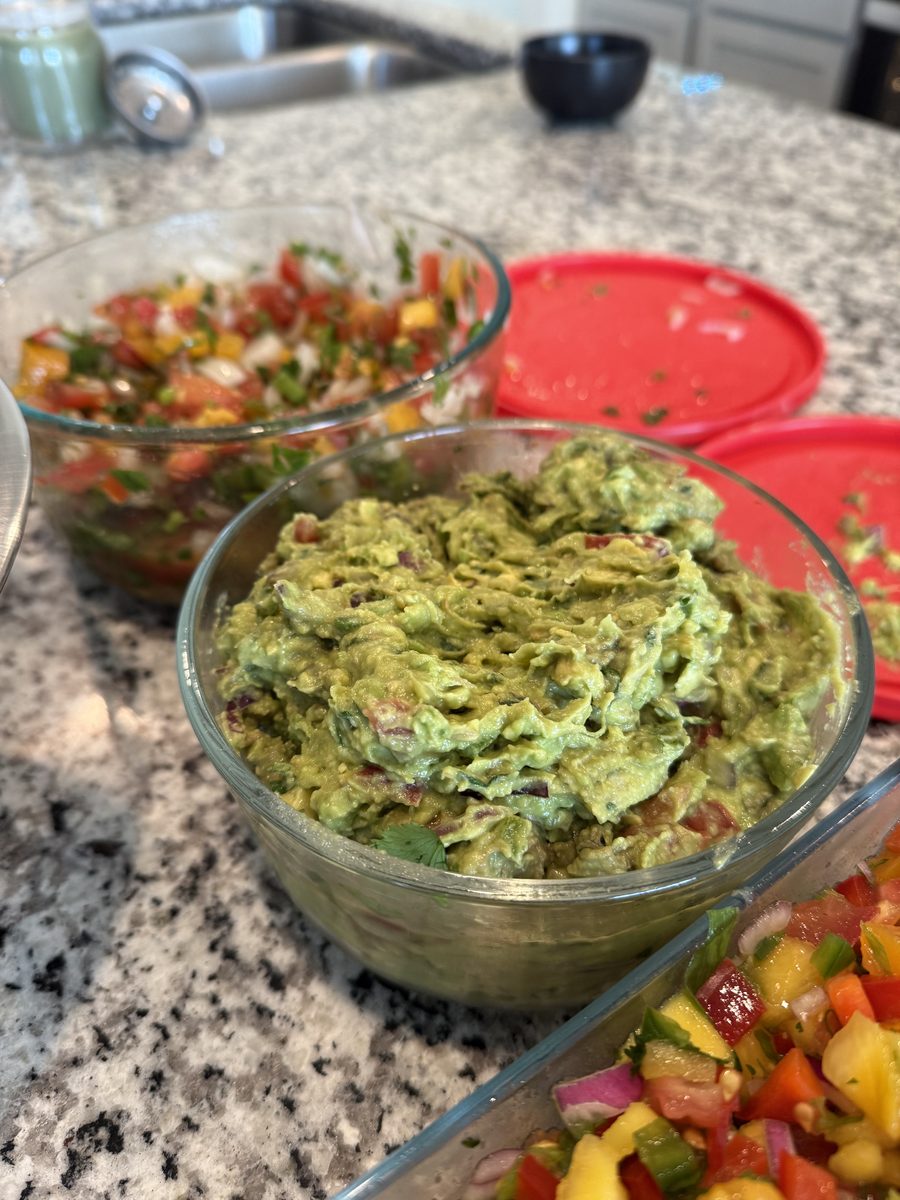 A glass bowl of chunky guacamole beside a bowl of pico de gallo and a tray of chopped peppers on a granite counter with a red stor