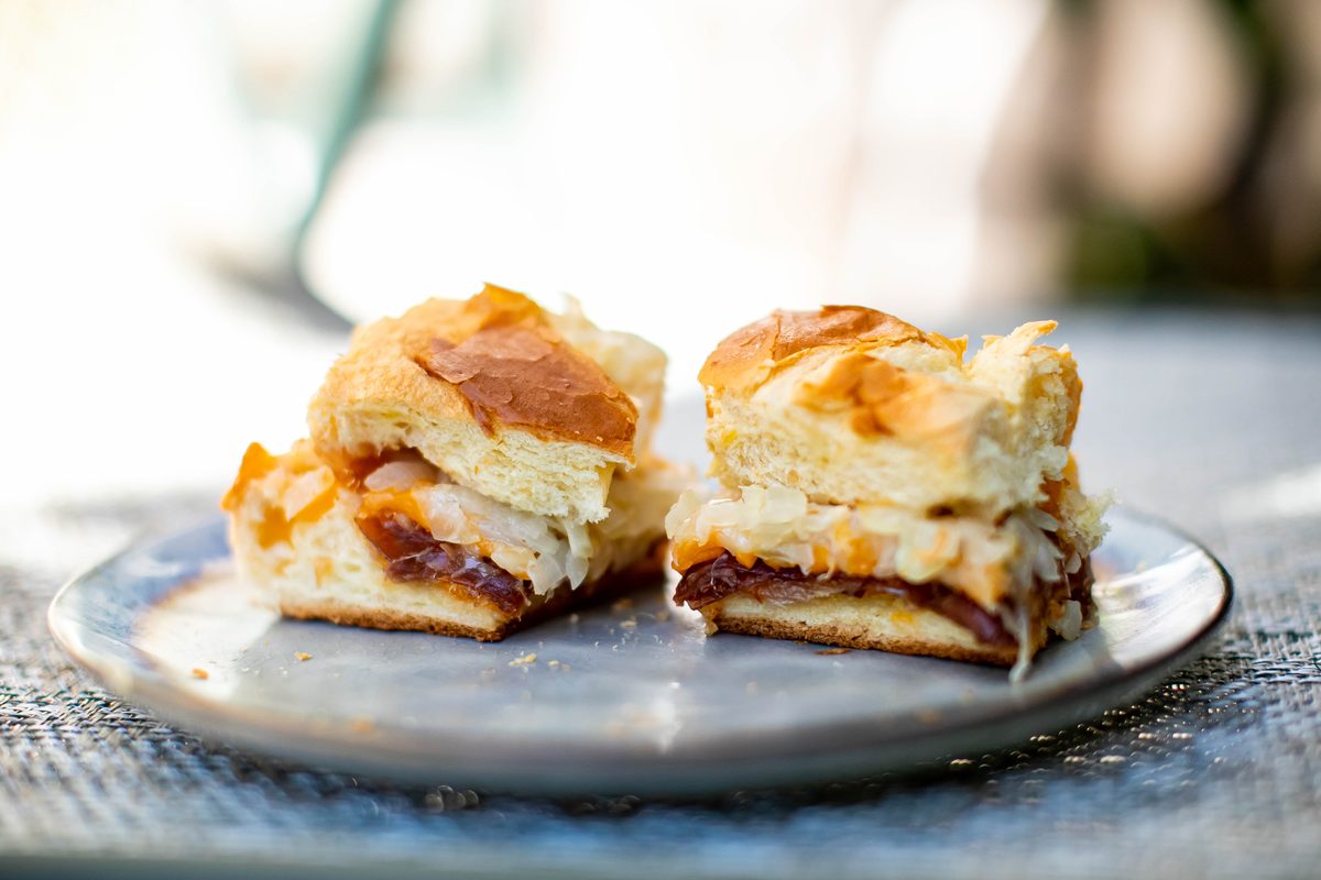 A grey plate holding two halved baked slider sandwiches on soft golden buns, layers of ham and melted cheese visible in the cut fa