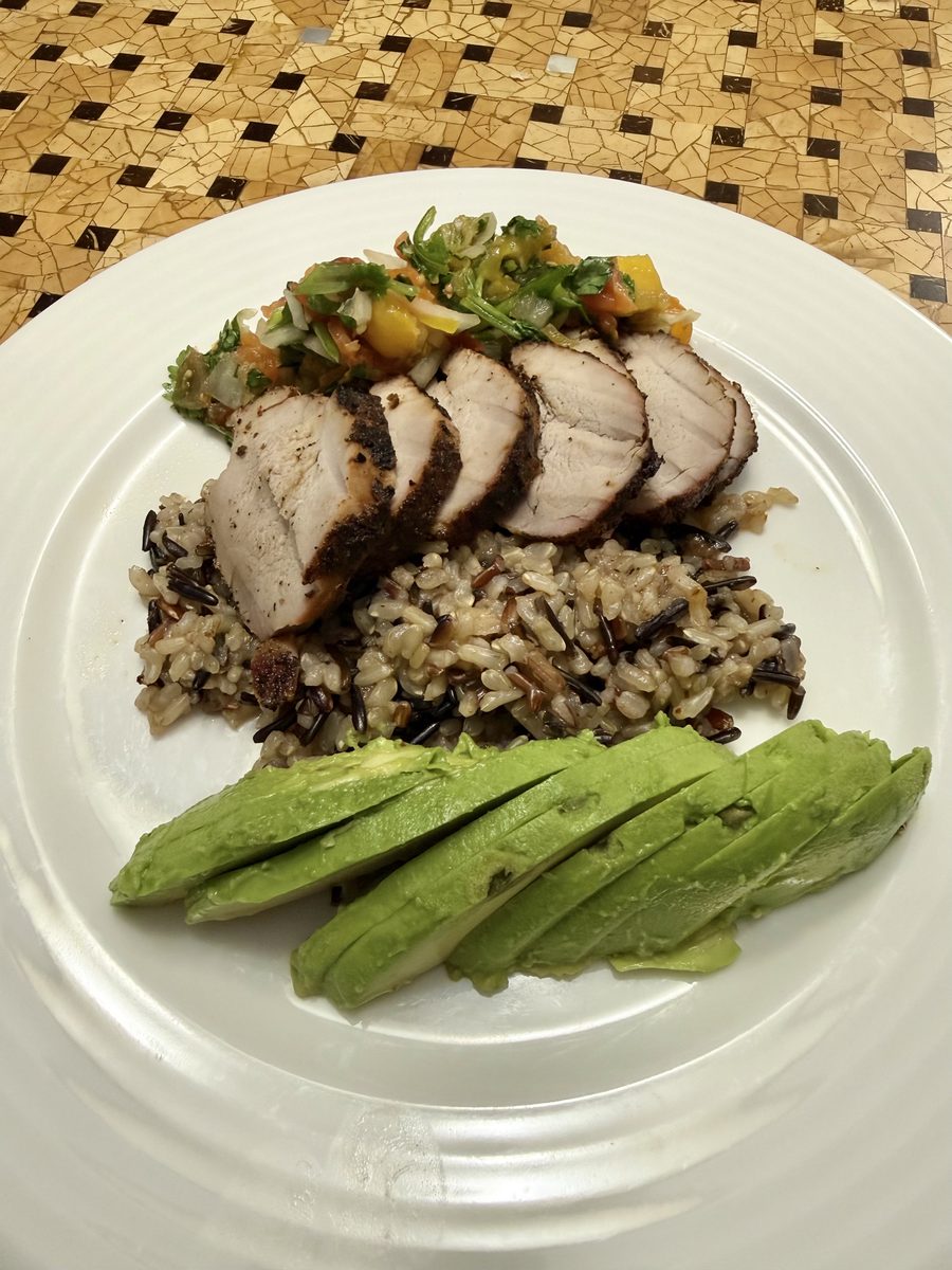 A white plate holding slices of herb-crusted roast pork topped with a chunky relish, a mound of wild rice pilaf and fanned avocado