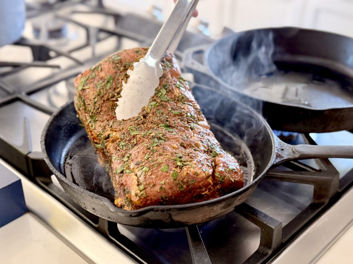 A herb-crusted pork loin roast being lifted with tongs in a cast iron skillet on a gas stovetop, a second smoking skillet beside i