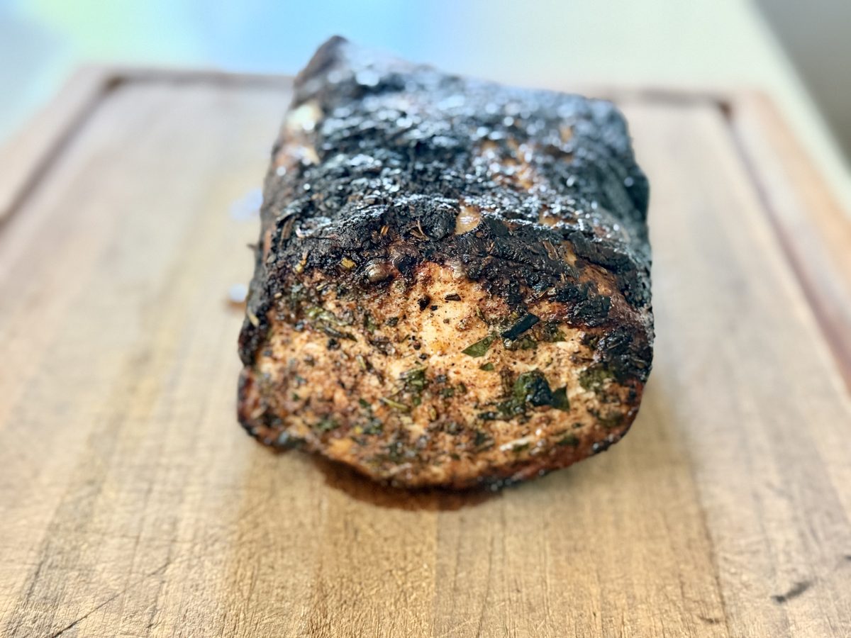 A whole roasted herb-crusted pork loin with a dark charred crust resting uncut on a wooden cutting board.