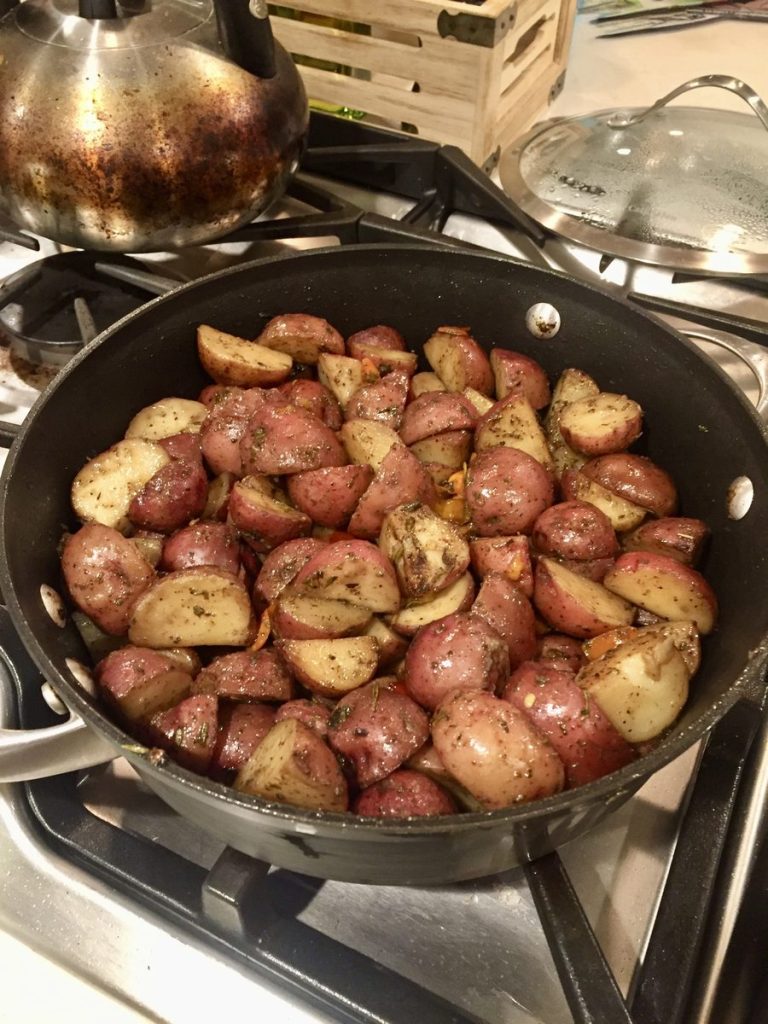 Browned halved baby red potatoes with herbs in a dark skillet on a gas stovetop, with a hand lifting the metal lid away above the pan.