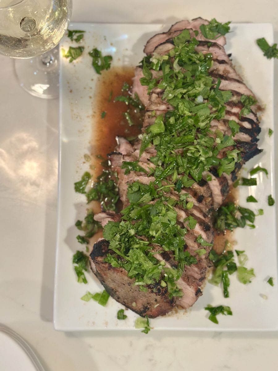 A long white rectangular platter with a row of thinly sliced pink roasted meat covered in chopped green herbs and pooled pan juice