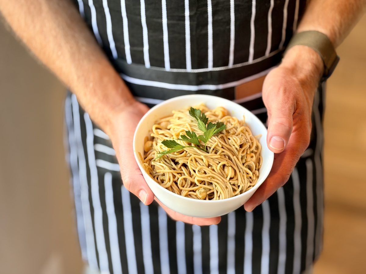 A man in a black-and-white striped apron holding a white bowl of spaghetti tossed with chickpeas and topped with a parsley sprig.