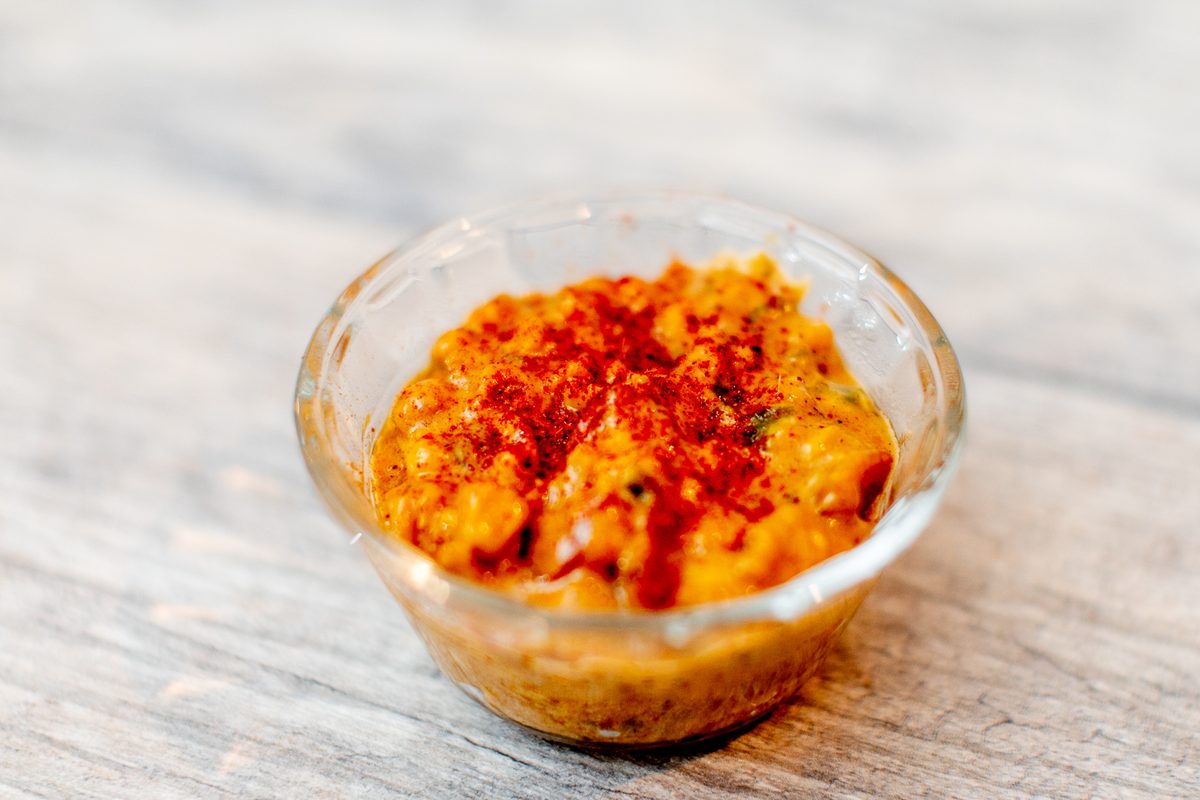 A clear glass bowl of creamy corn dusted heavily with red paprika, on a pale wooden table.