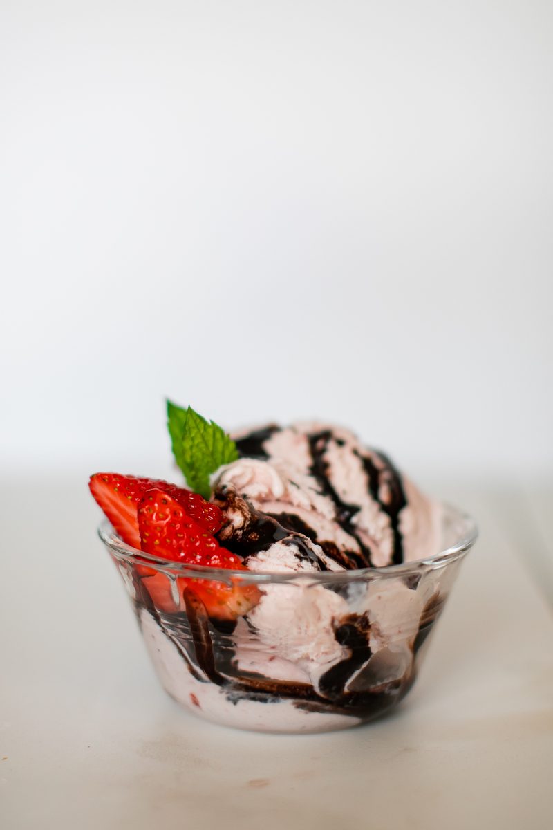 A glass dessert bowl of pale pink ice cream drizzled with chocolate sauce and garnished with a sliced strawberry and mint, on a wh