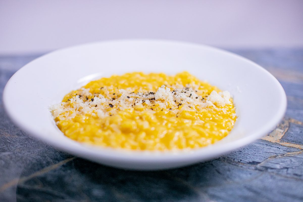 A wide white bowl of bright yellow butternut squash risotto topped with grated parmesan and cracked pepper, on a blue marble table