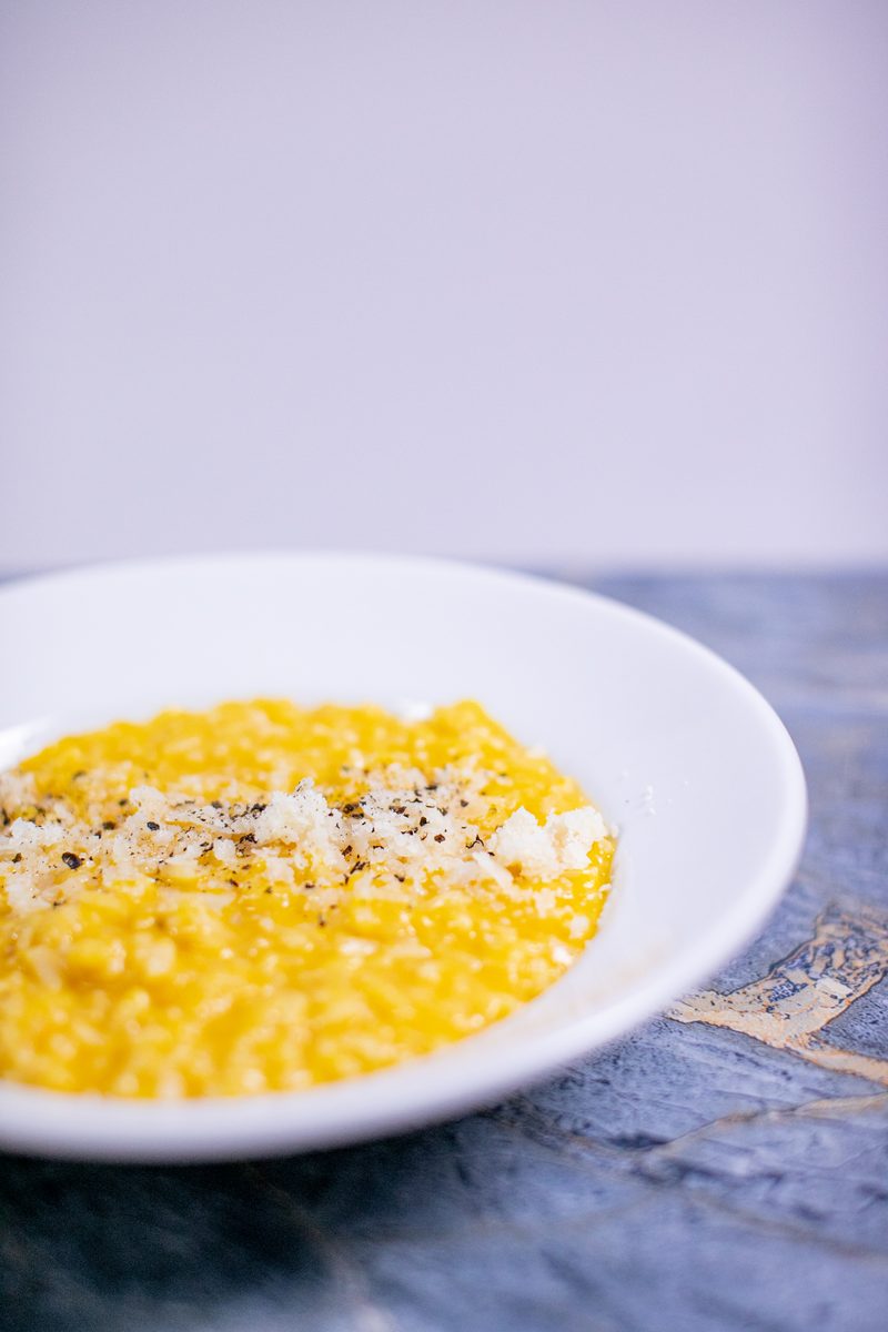 Tighter crop of the same white bowl of yellow butternut squash risotto with parmesan and black pepper on a blue marble surface.