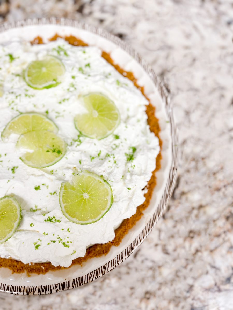 An overhead view of a whole key lime pie in a glass dish, topped with whipped cream and a ring of lime slices, on a speckled stone