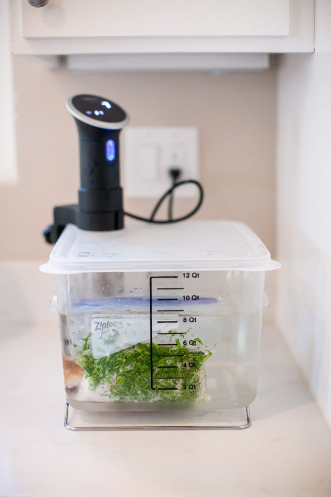 A black sous vide immersion circulator clipped to a clear plastic water bath tub on a white counter, with a bag of herb-marinated chicken submerged inside.