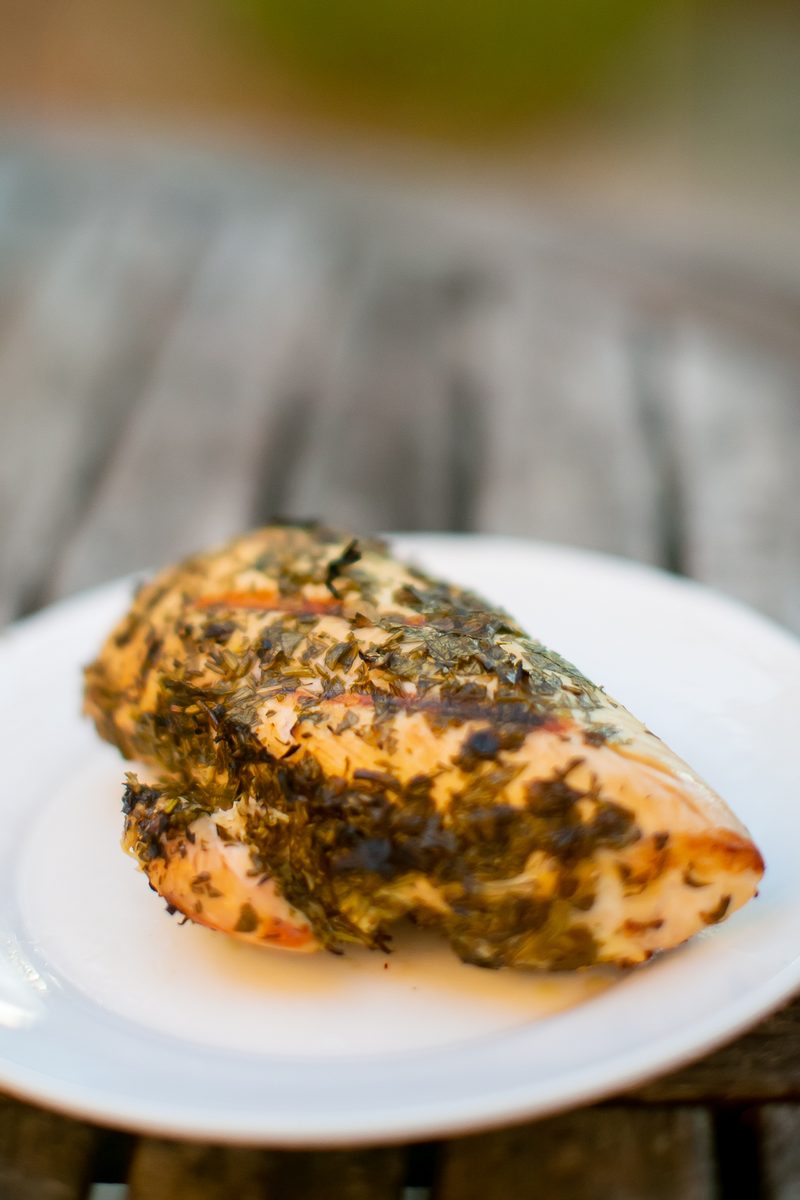 A cooked herb-crusted chicken breast plated on a white dish with pan juices pooled around it, on a weathered wood table outdoors.