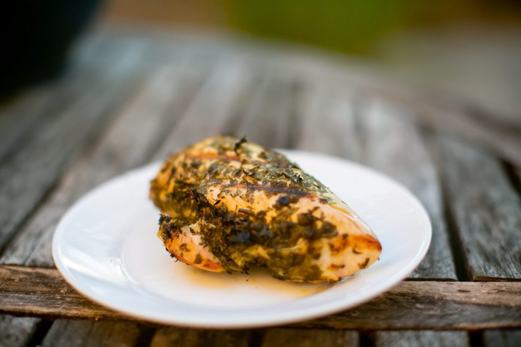 Close view of a golden herb-topped chicken breast on a white plate with juices, set on rustic grey wood planks.