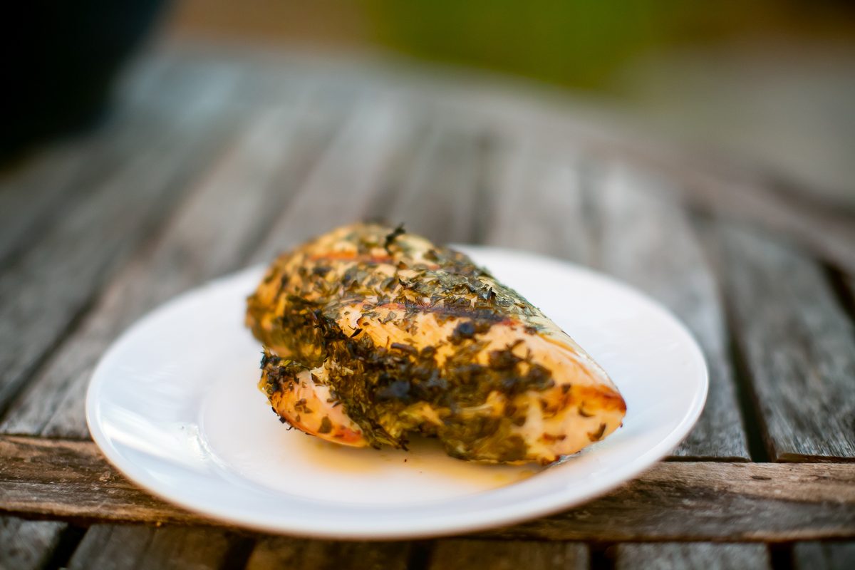 Close view of a golden herb-topped chicken breast on a white plate with juices, set on rustic grey wood planks.