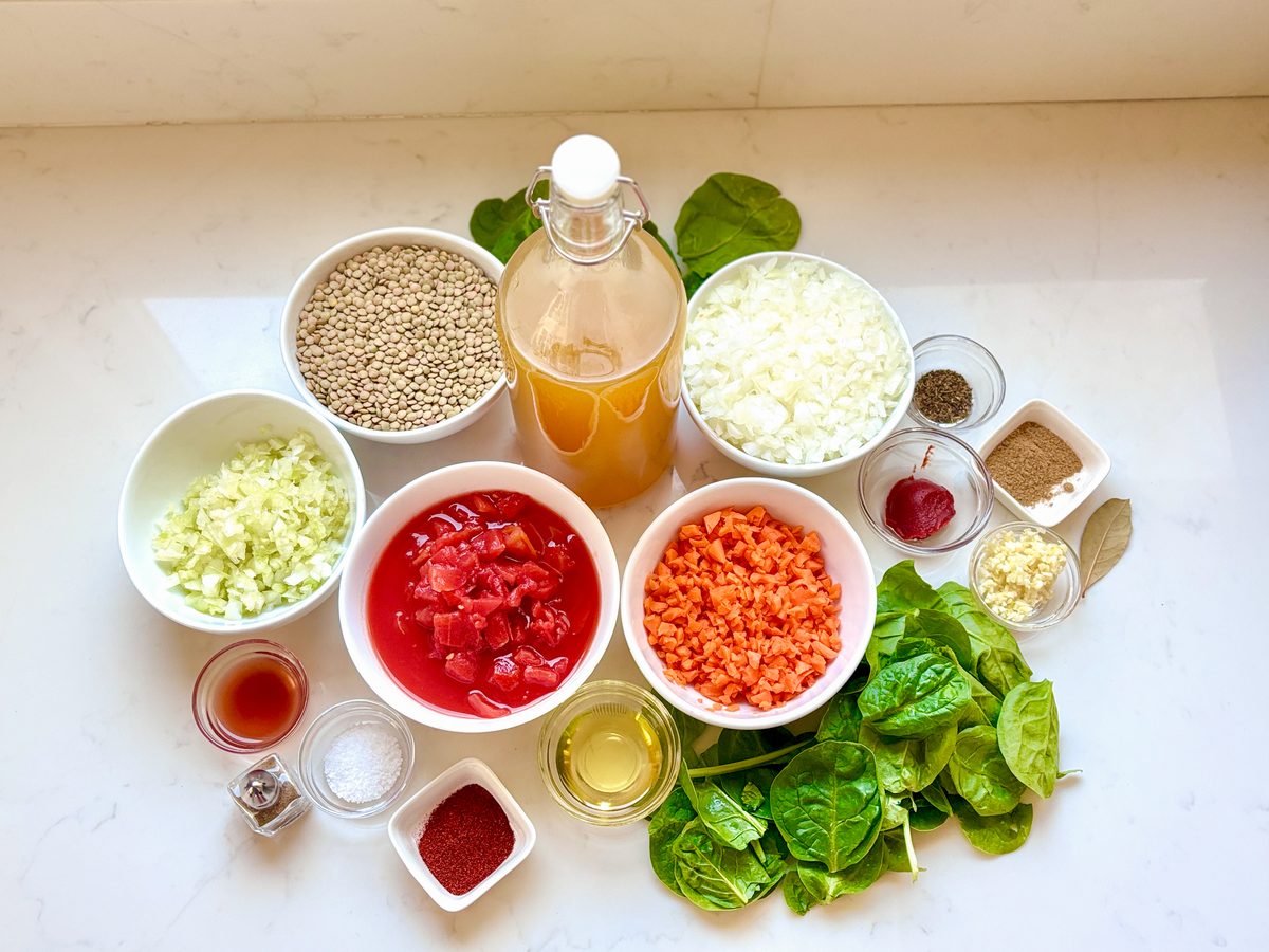 An overhead flat lay of lentil soup ingredients in white bowls - dried lentils, diced onion, canned tomatoes, chopped carrots, spi
