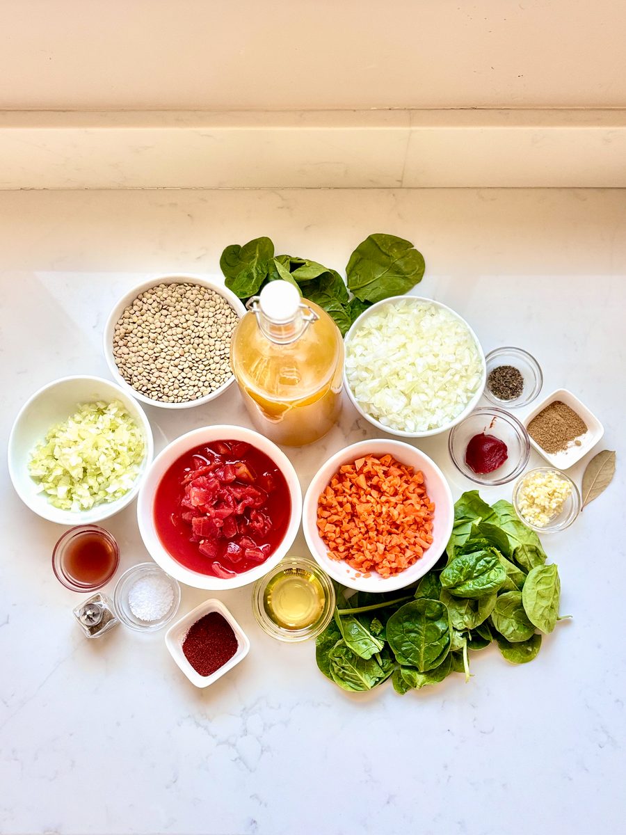 A tighter overhead of the same lentil soup ingredient spread with bowls of lentils, tomatoes, carrots, onion, spinach and oil on a
