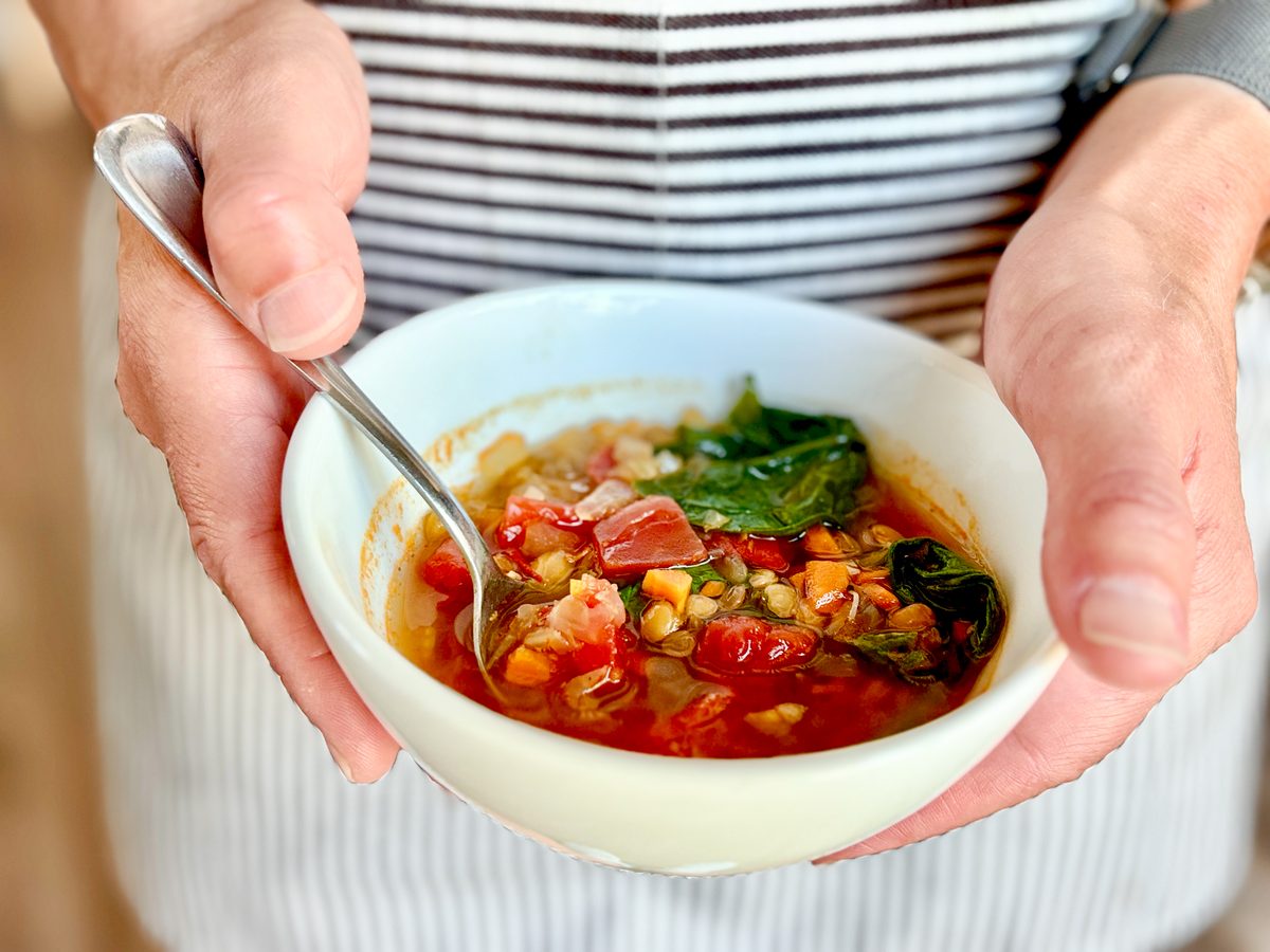 Two hands holding a white bowl of finished lentil and vegetable soup with a spoon in it, against a striped apron.