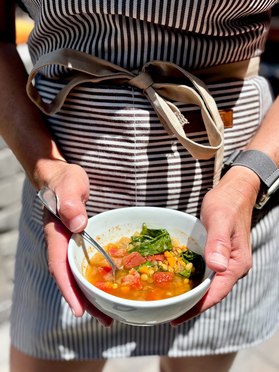 A person in a striped apron holding a white bowl of brothy vegetable soup with tomato, corn and greens and a spoon, shot in harsh