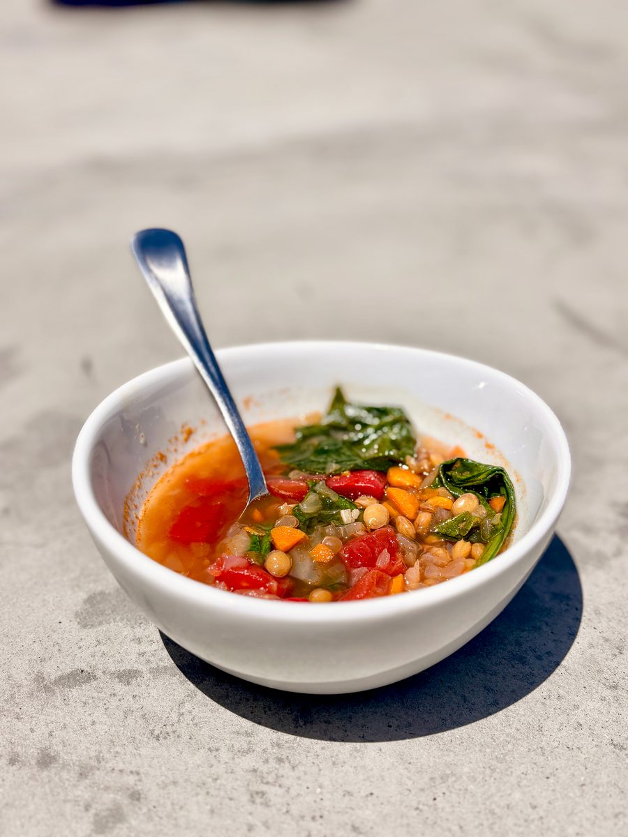A white bowl of tomato-based vegetable and lentil soup with spinach and a spoon resting in it, on a dark round mat over a light su