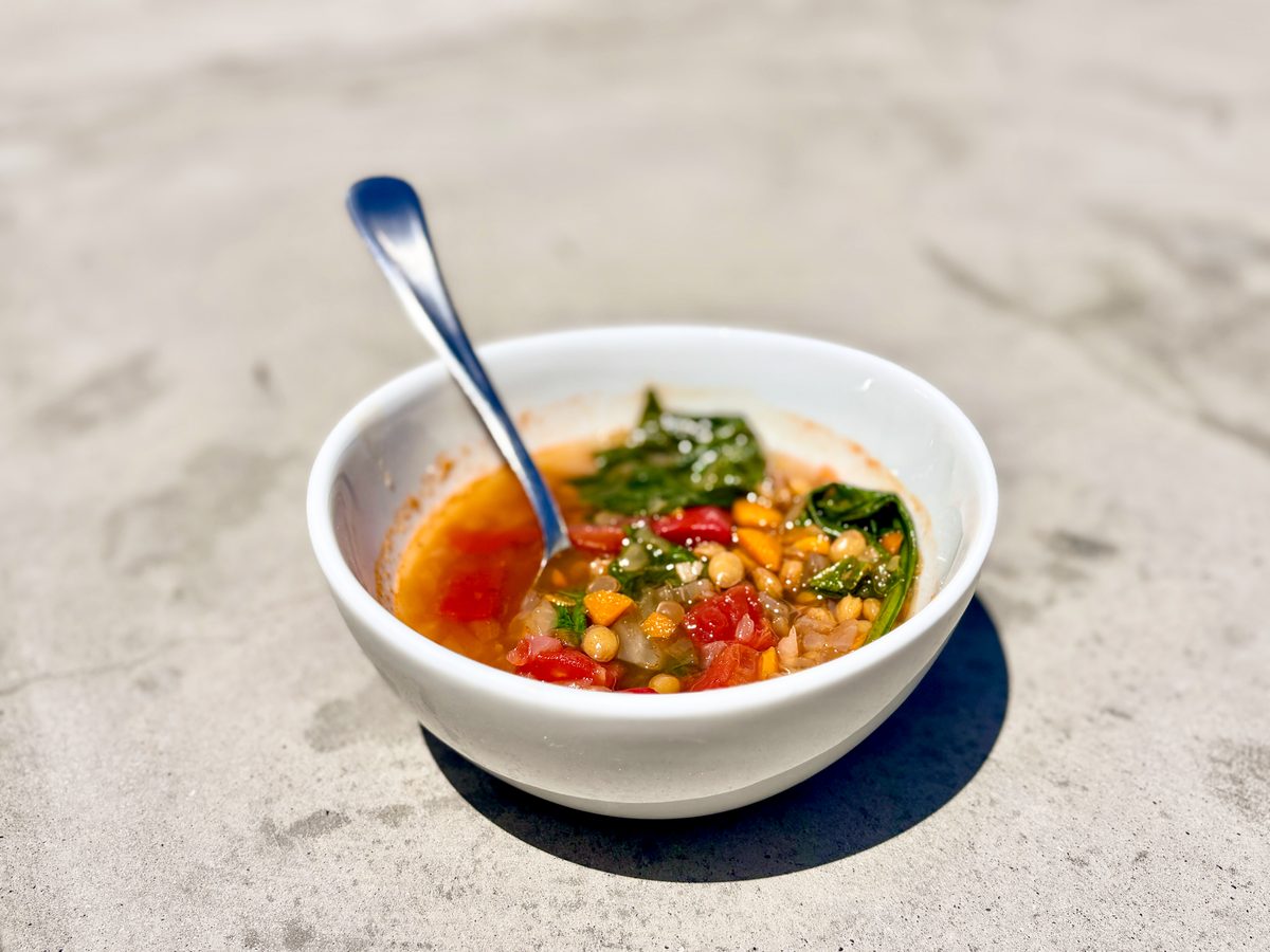 A white bowl of chunky vegetable lentil soup with tomato, corn and spinach, spoon in the bowl, on a grey concrete surface.