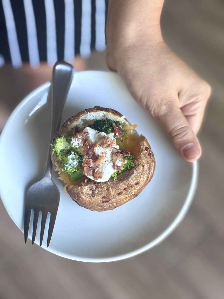 Two hands holding a pale blue plate with a finished loaded baked potato topped with broccoli, cheese, sour cream and bacon, plus a