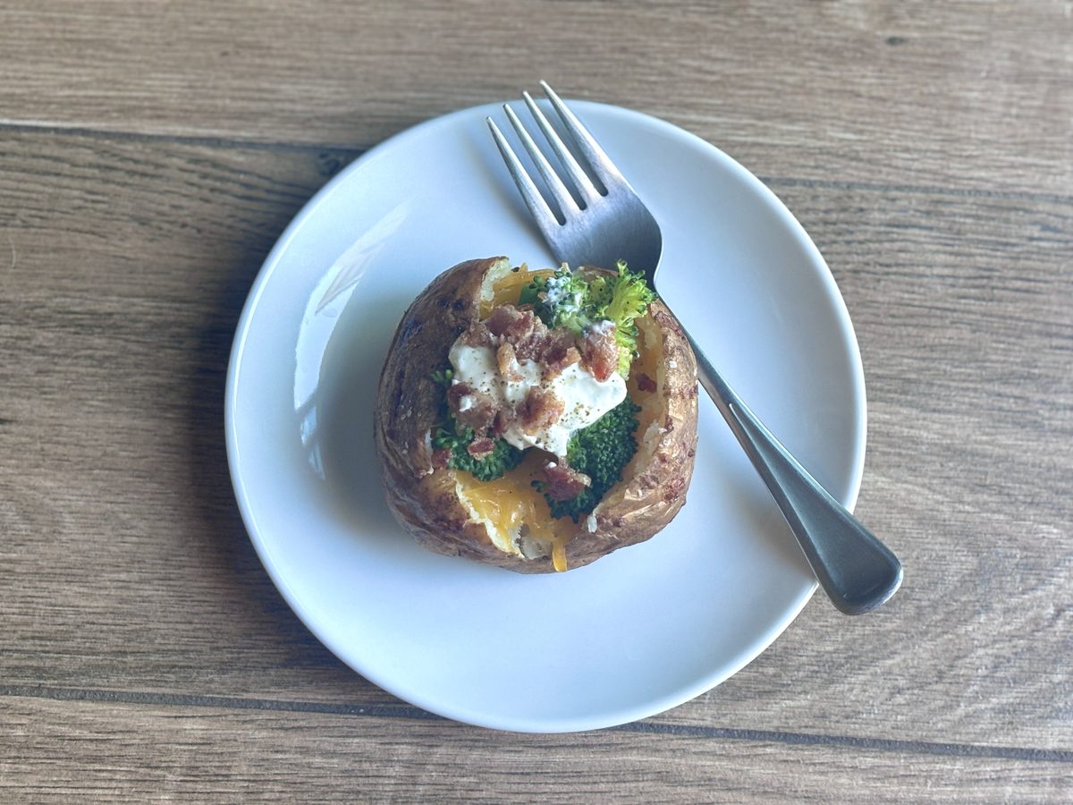Overhead shot of a loaded baked potato with broccoli, cheddar, sour cream and bacon on a white plate with a fork, on a grey weathe