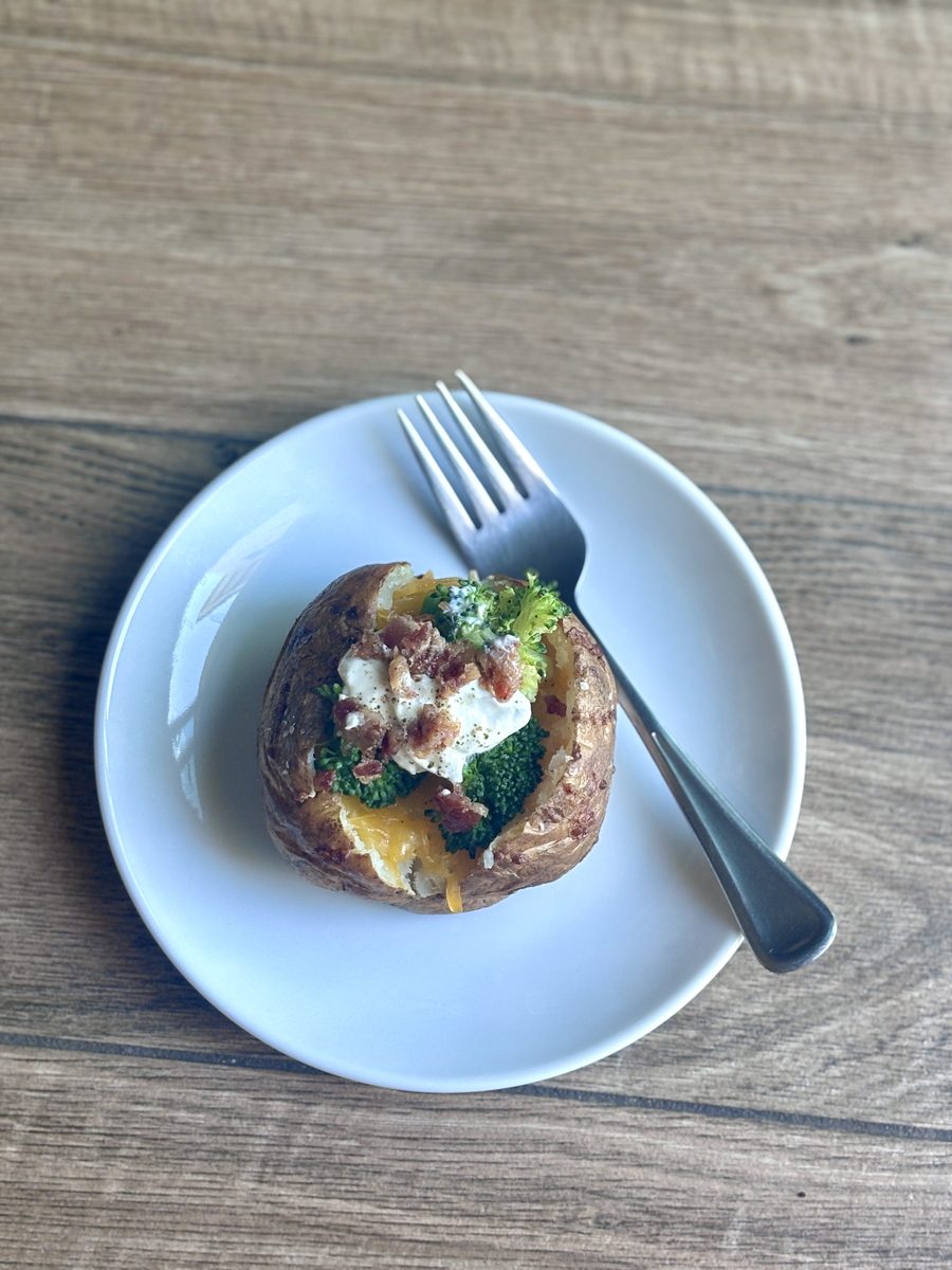 A finished loaded baked potato with broccoli, cheese, sour cream and bacon served on a pale blue plate with a fork on weathered wo