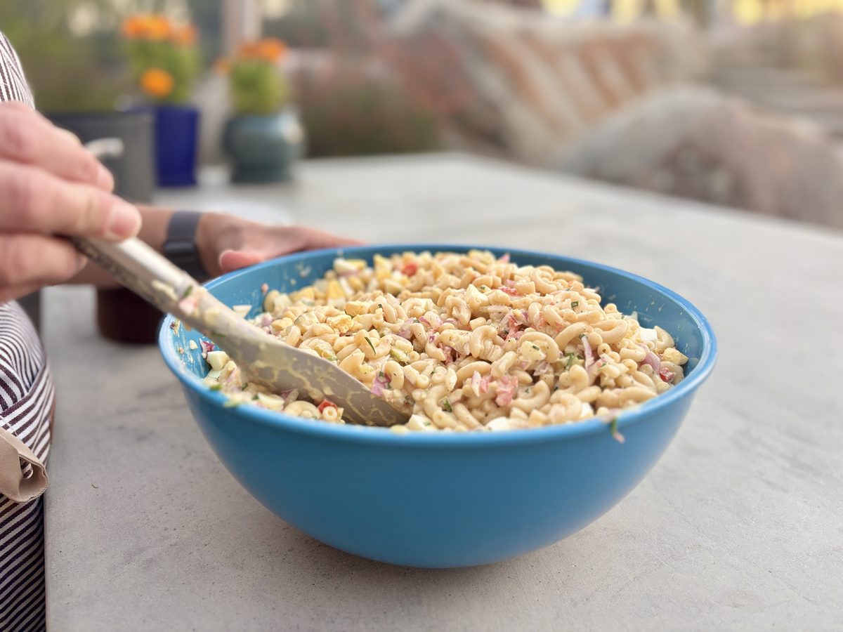 A blue ceramic serving bowl of creamy macaroni salad with pink and green flecks on an outdoor stone patio table, a hand resting a